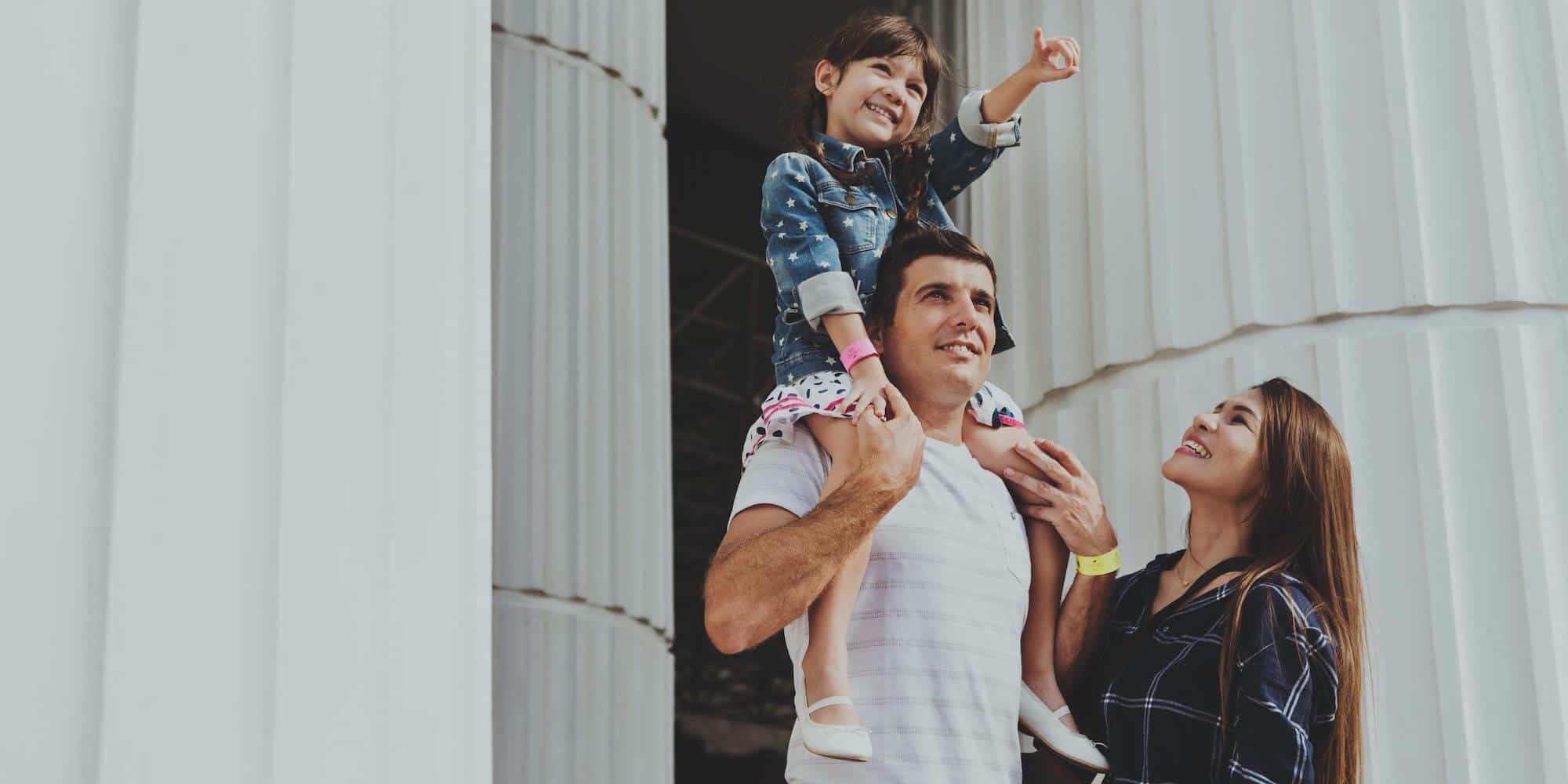 A smiling girl on her dad's shoulders, pointing to the sky, with her mum beside them